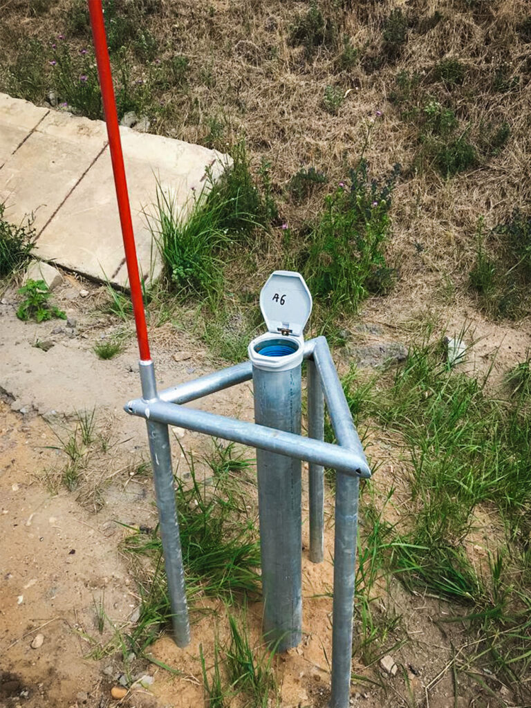 Nahaufnahme einer oberirdisch ausgebauten Grundwassermessstelle im Außenbereich. Zu sehen ist ein senkrechtes, graues Metallrohr, das aus dem sandig-grasigen Boden ragt.