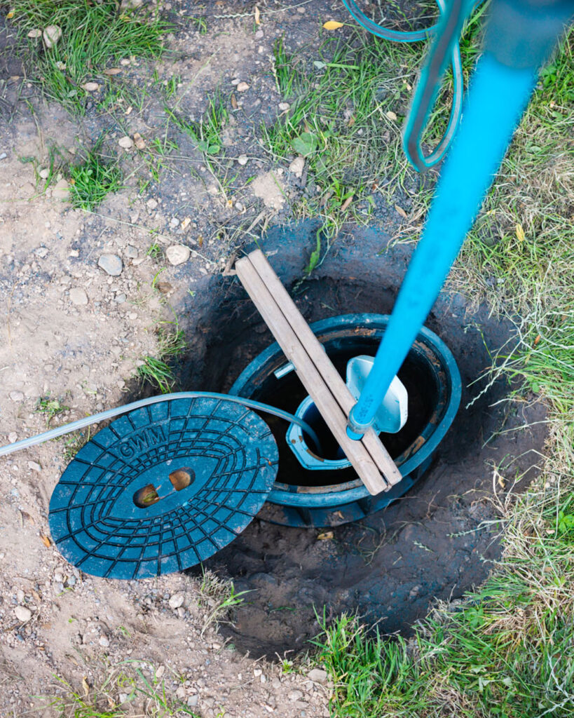 Blick von oben in einen tiefer liegenden, geöffneten Grundwassermessschacht im Erdreich. Der ovale, gusseiserne Verschlussdeckel mit der Aufschrift „GWM“ liegt links daneben im Gras.